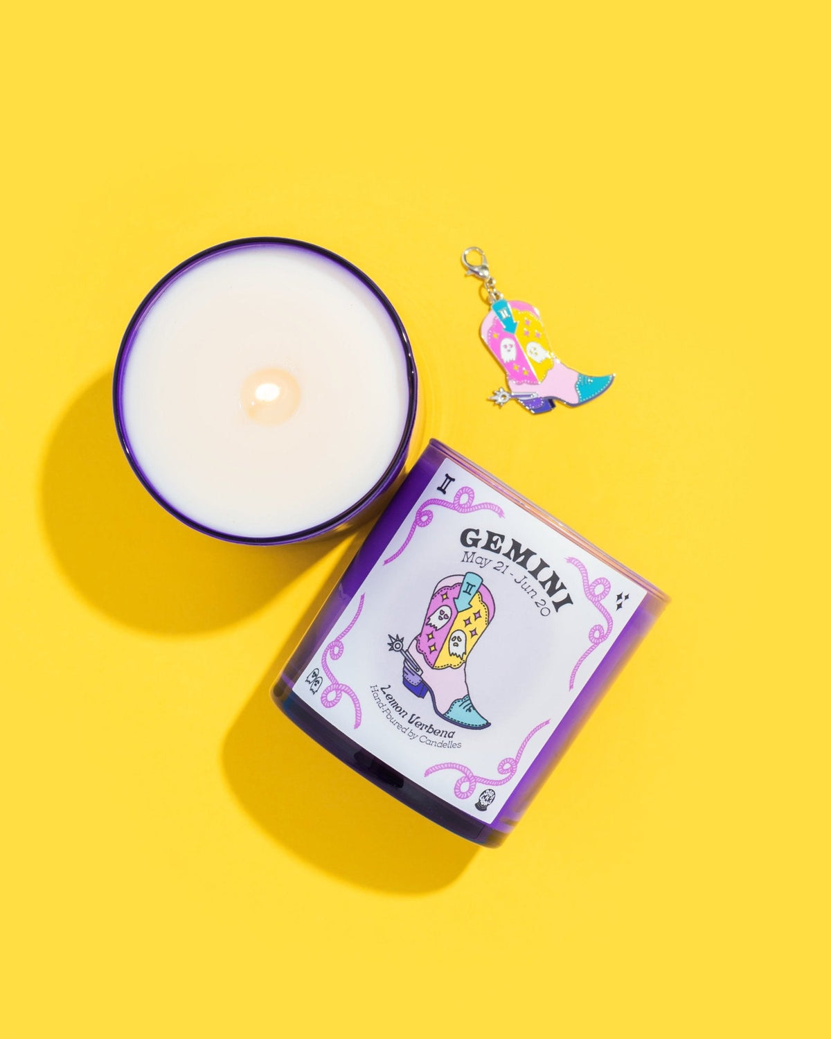Lemon verbena Gemini zodiac candle beside a lit purple glass jar and a cowboy boot charm on a yellow background