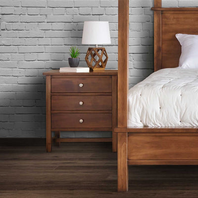 Three-drawer bedside chest in warm brown wood with nickel knobs beside a canopy bed post, lamp and books against white brick