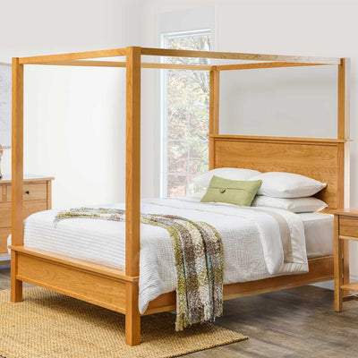 Natural cherry canopy queen bed with white quilted bedding and a knit throw beside a window in a bright bedroom