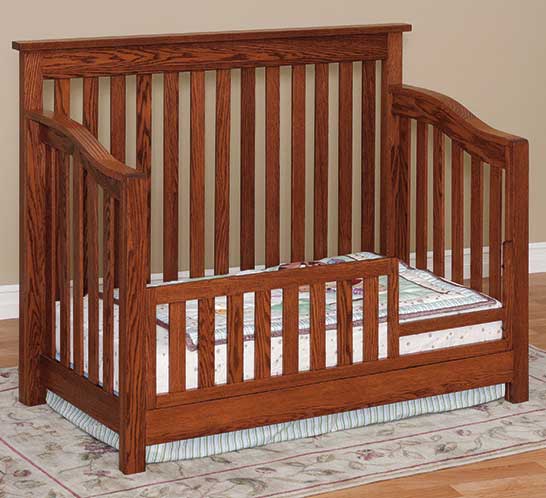Mission style oak crib with straight slats on all sides, curved top rails and pastel crib bedding beside a floor lamp
