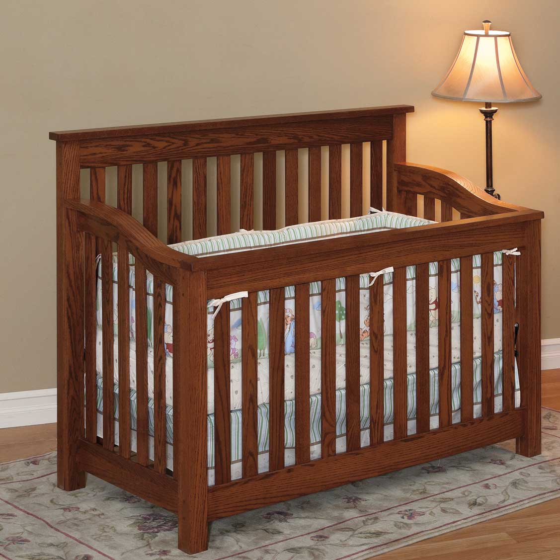 Mission style oak crib with straight slats on all sides, curved top rails and pastel crib bedding beside a floor lamp