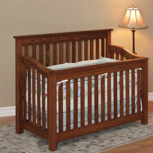Mission style oak crib with straight slats on all sides, curved top rails and pastel crib bedding beside a floor lamp