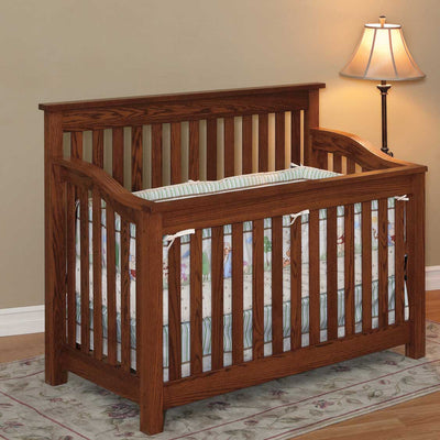 Mission style oak crib with straight slats on all sides, curved top rails and pastel crib bedding beside a floor lamp
