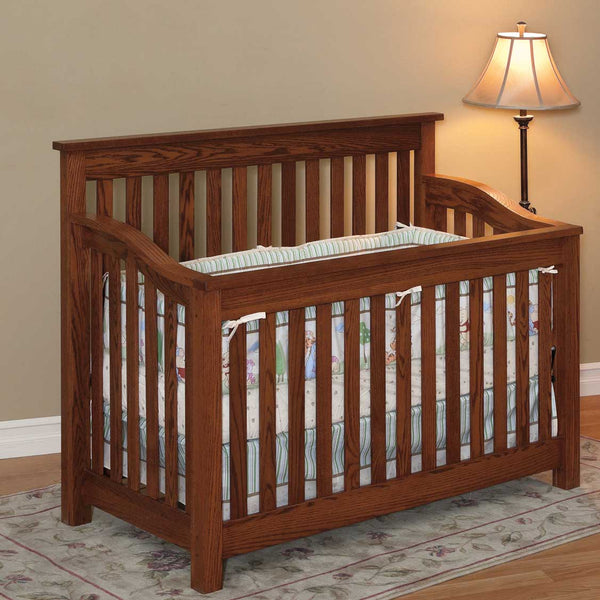 Mission style oak crib with straight slats on all sides, curved top rails and pastel crib bedding beside a floor lamp