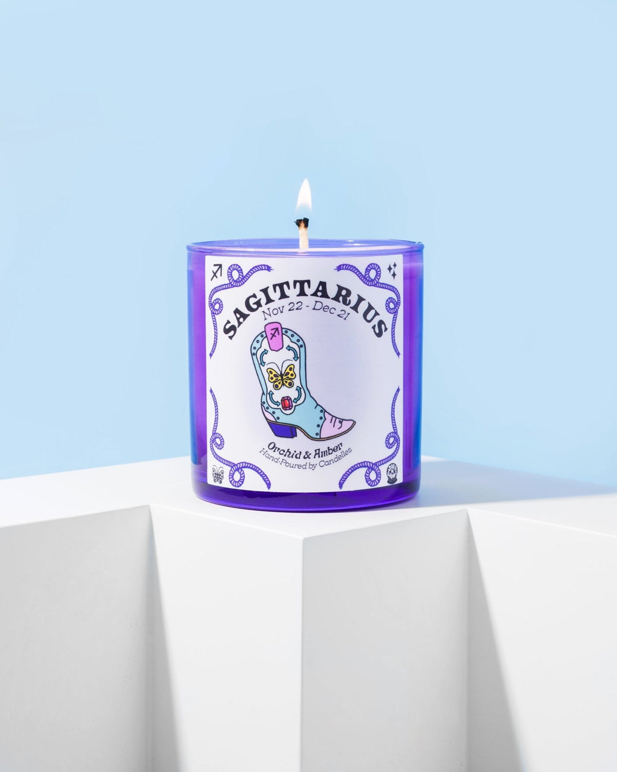 Orchid and amber Sagittarius zodiac candle burning in a purple glass tumbler on a white block against pale blue