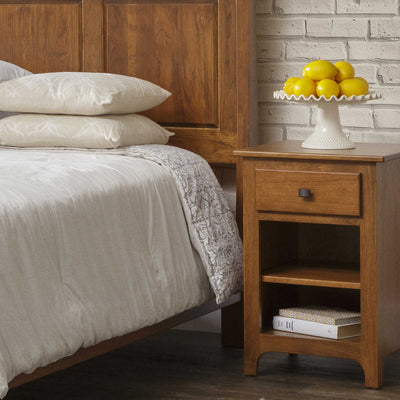 Small cherry open nightstand with one drawer, black bar pull and two open shelves holding books beside a bed