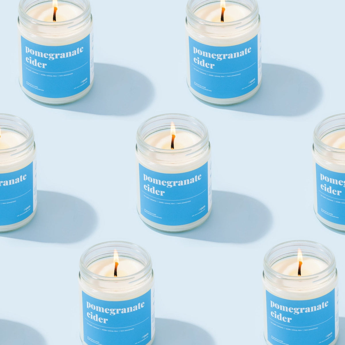 Repeating grid of lit pomegranate cider soy candles with blue labels arranged across a pale blue background