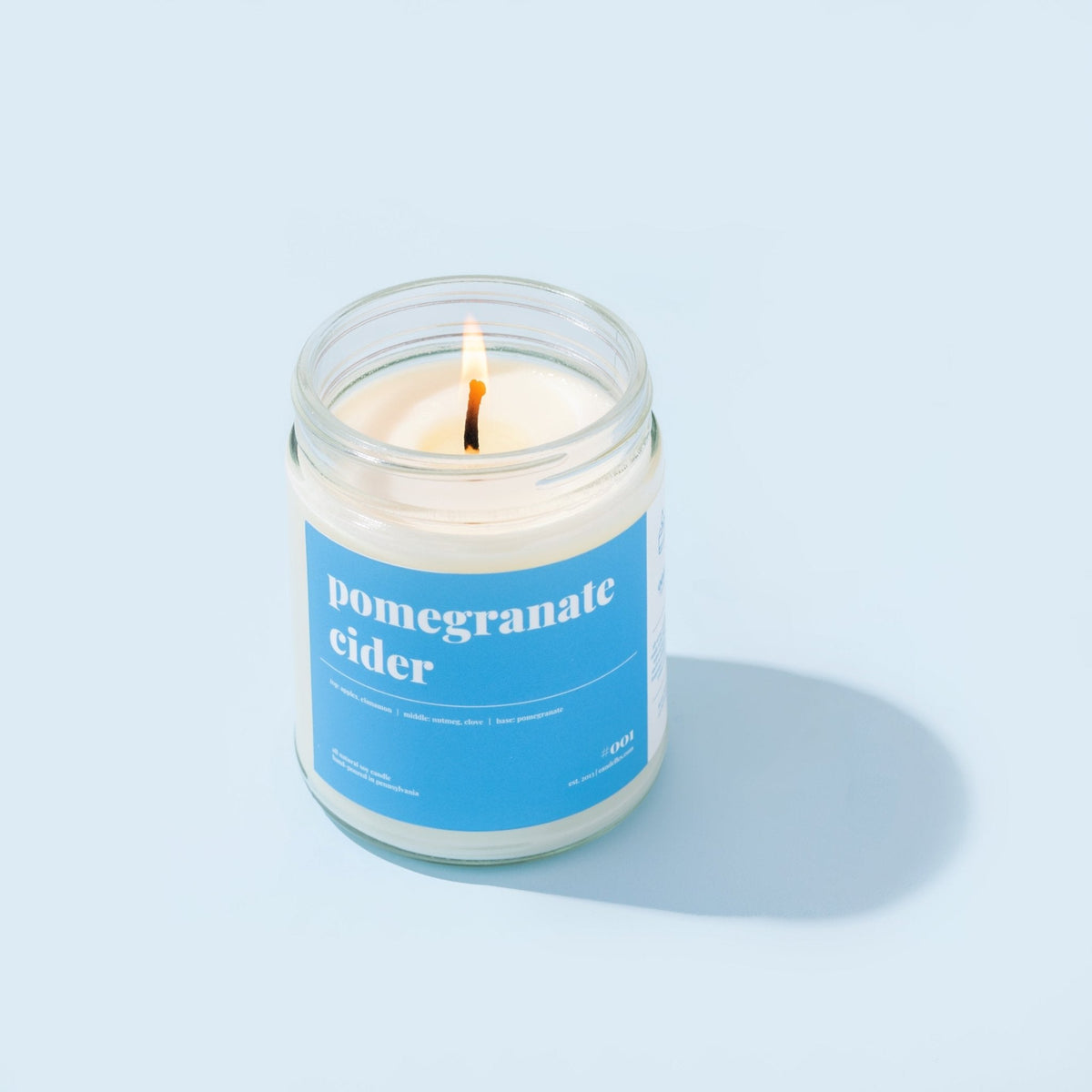 Pomegranate cider soy candle burning in a clear jar with a bright blue label on a pale blue background