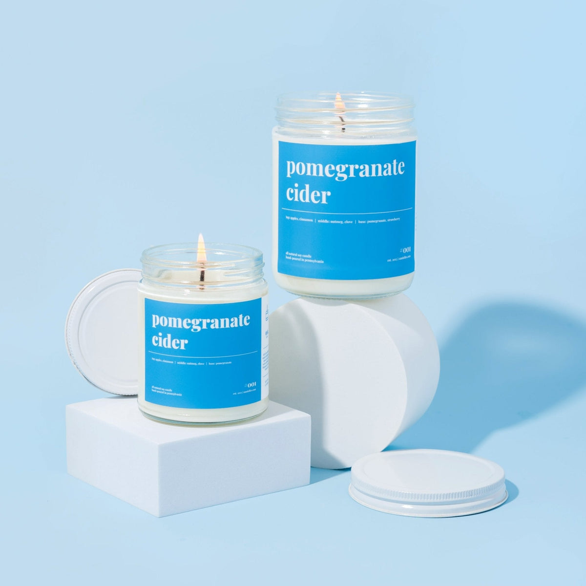 Two pomegranate cider soy candles lit on white risers with loose lids against a soft blue background
