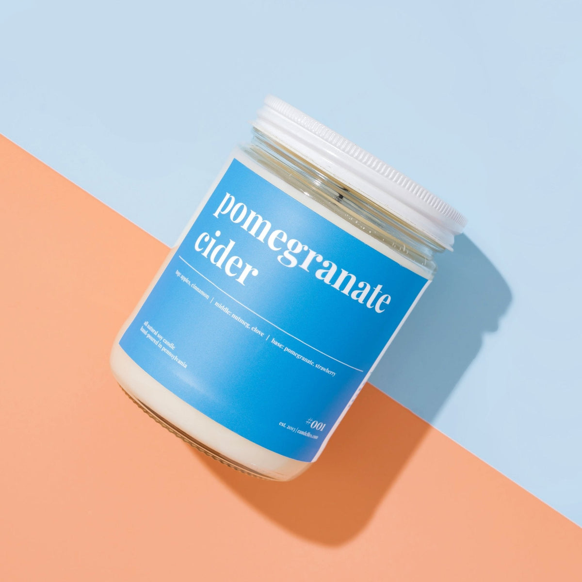Overhead lidded pomegranate cider soy candle with blue label on a split blue and peach paper background