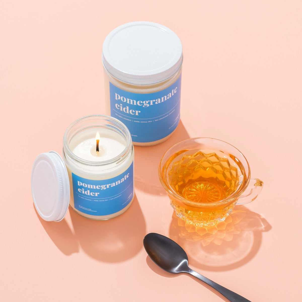 Two pomegranate cider soy candles, one lit with lid off, beside a glass teacup of amber cider on peach