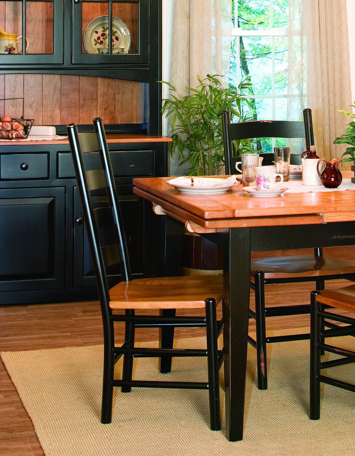 Black ladder back chairs with cherry seats pulled up to a two-tone draw leaf dining table in a country kitchen