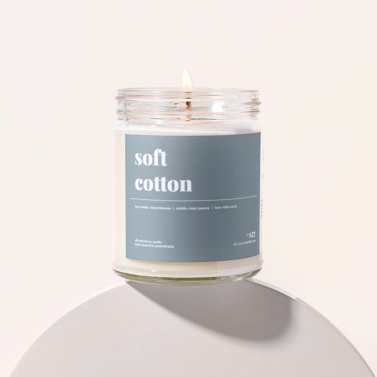 Soft cotton soy candle burning in a clear jar with a slate blue label on a white pedestal against cream