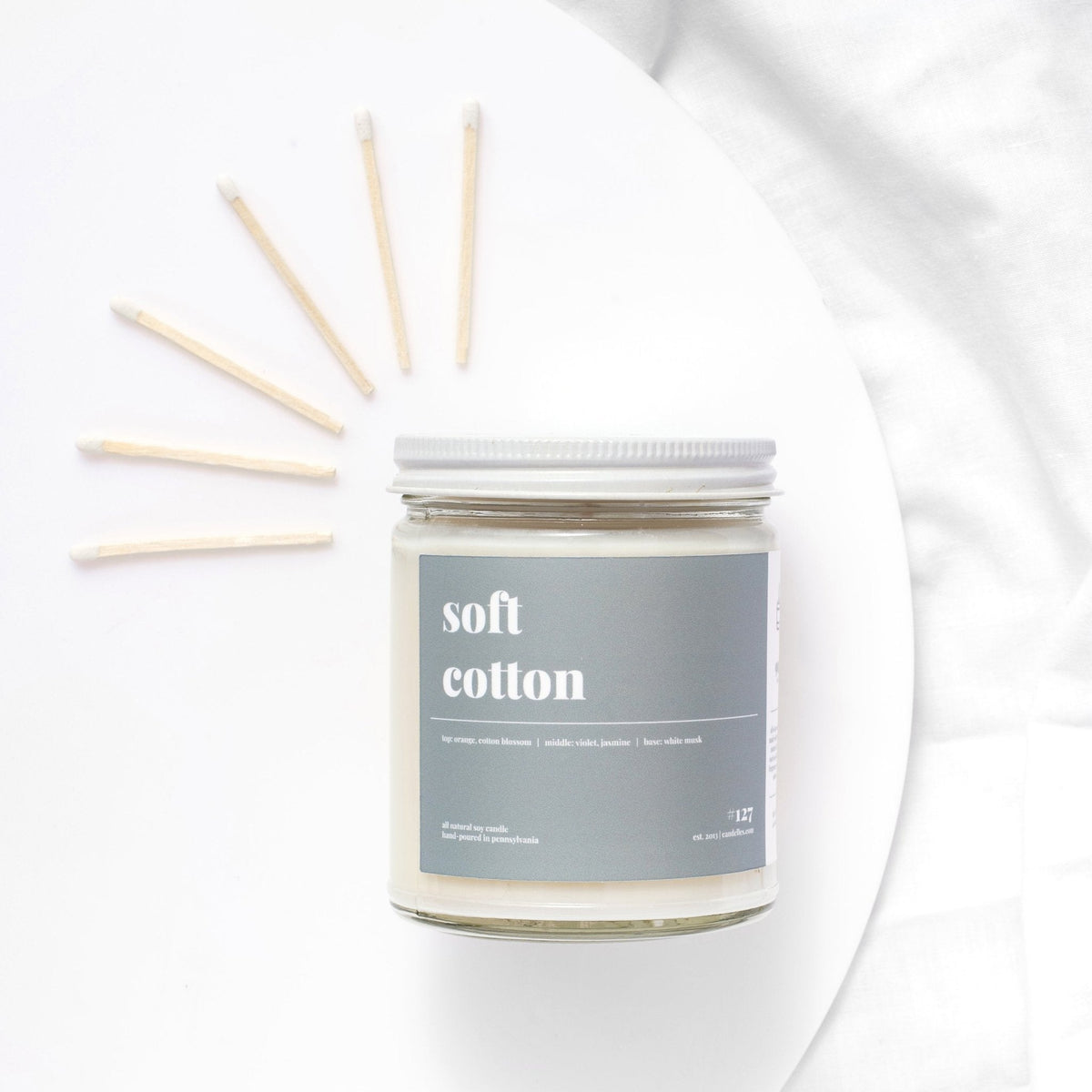 Soft cotton soy candle with white lid on a white plate with matches fanned out behind it on white linen