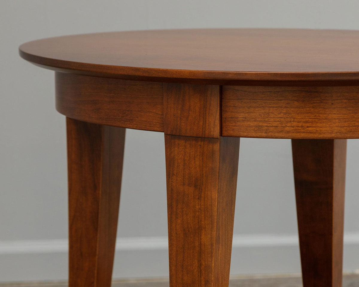 Angled detail of the round table top, apron and three tapered legs in a glossy cherry finish