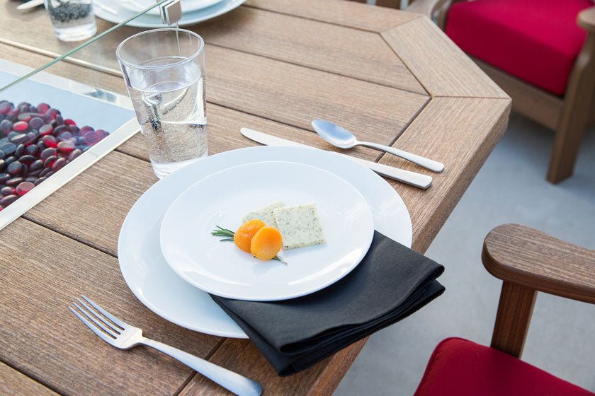 Place setting with white plates and black napkin on a teak-toned poly fire table beside red cushioned chairs