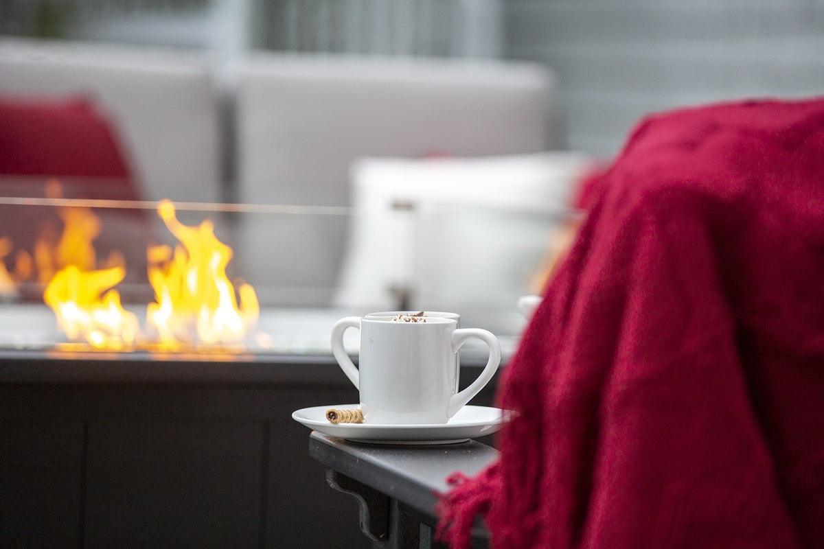 White cup of cocoa and a red throw blanket beside the low flames of a lit outdoor fire table