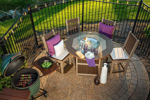 Aerial view of a round fire pit counter table with glass guard, four cushioned bar chairs and grilled kabobs on a nearby ceramic grill