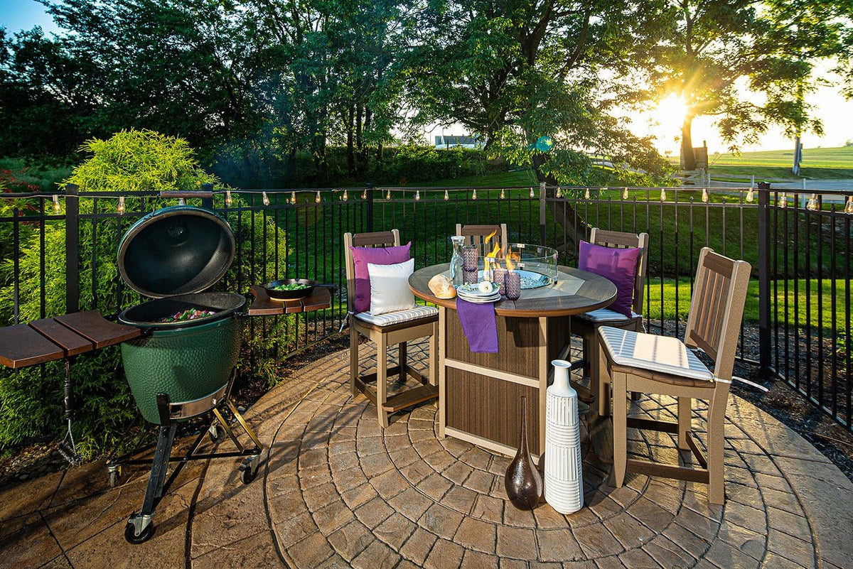 Backyard patio scene with a lit poly fire pit counter table, four bar chairs and a green ceramic grill under string lights at sunset