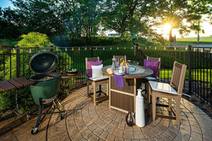 Backyard patio scene with a lit poly fire pit counter table, four bar chairs and a green ceramic grill under string lights at sunset