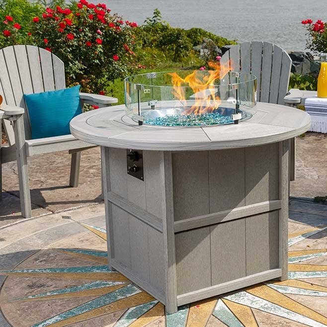 Gray poly round fire pit dining table with paneled base and blue glass beads burning under a clear glass guard on a decorative patio