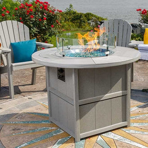 Gray poly round fire pit dining table with paneled base and blue glass beads burning under a clear glass guard on a decorative patio