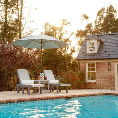 Pair of gray poly sun loungers with white cushions sharing a side table and green market umbrella beside a pool