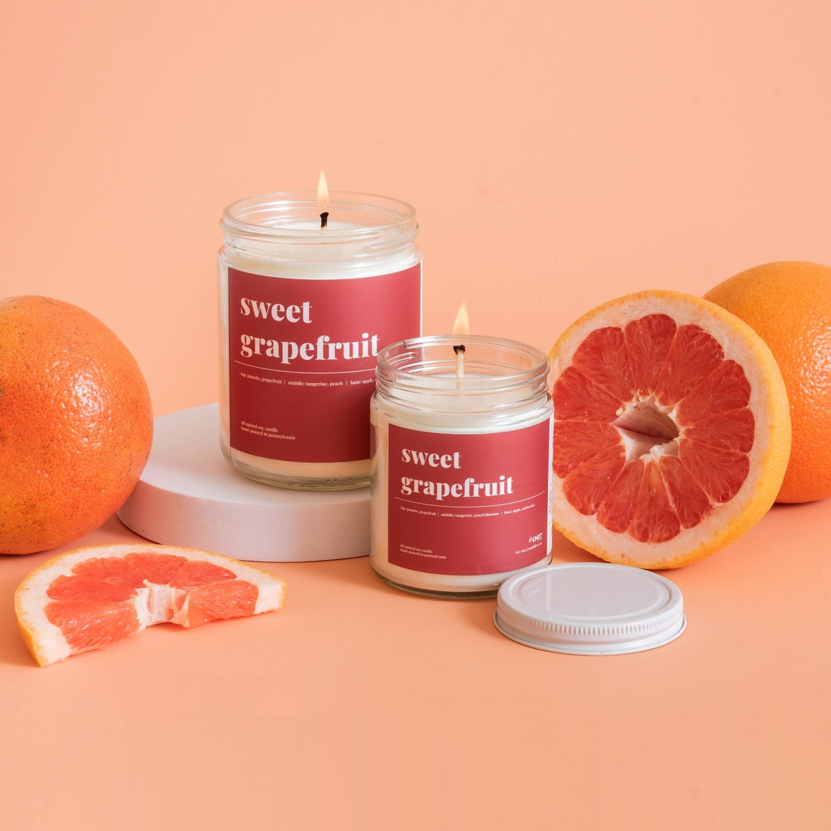 Two lit sweet grapefruit soy candles beside whole and halved pink grapefruit on a peach backdrop