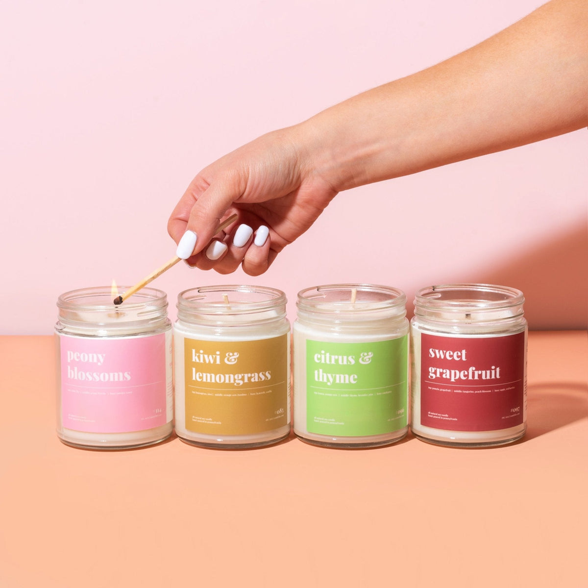 Hand lighting a row of four soy candles — peony blossoms, kiwi and lemongrass, citrus and thyme, sweet grapefruit