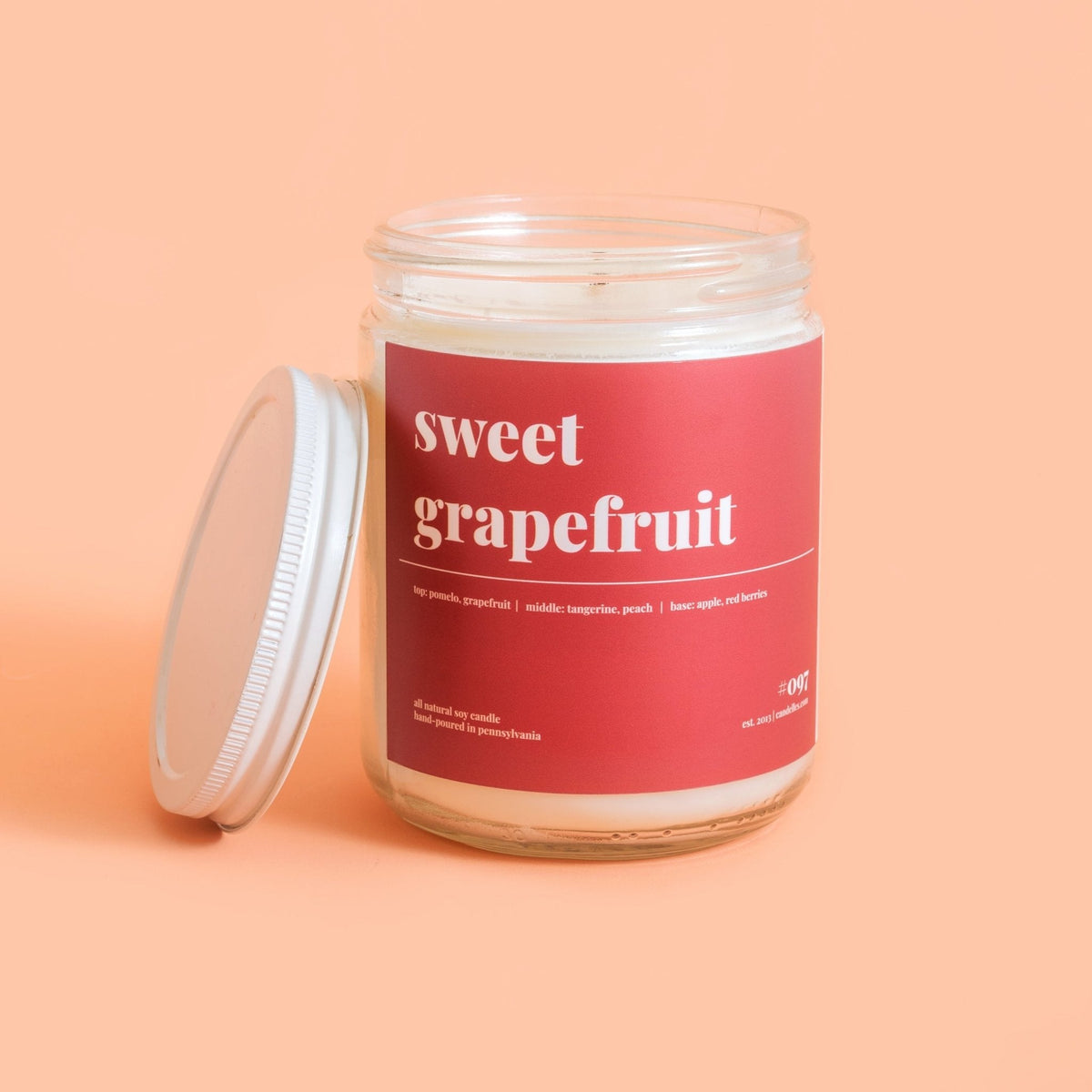 Sweet grapefruit soy candle with its white lid leaning against the jar on a warm peach background