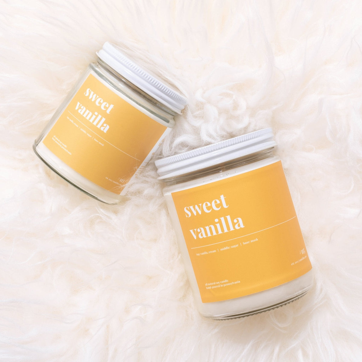 Overhead pair of sweet vanilla soy candles with white lids and golden labels resting on fluffy white fur