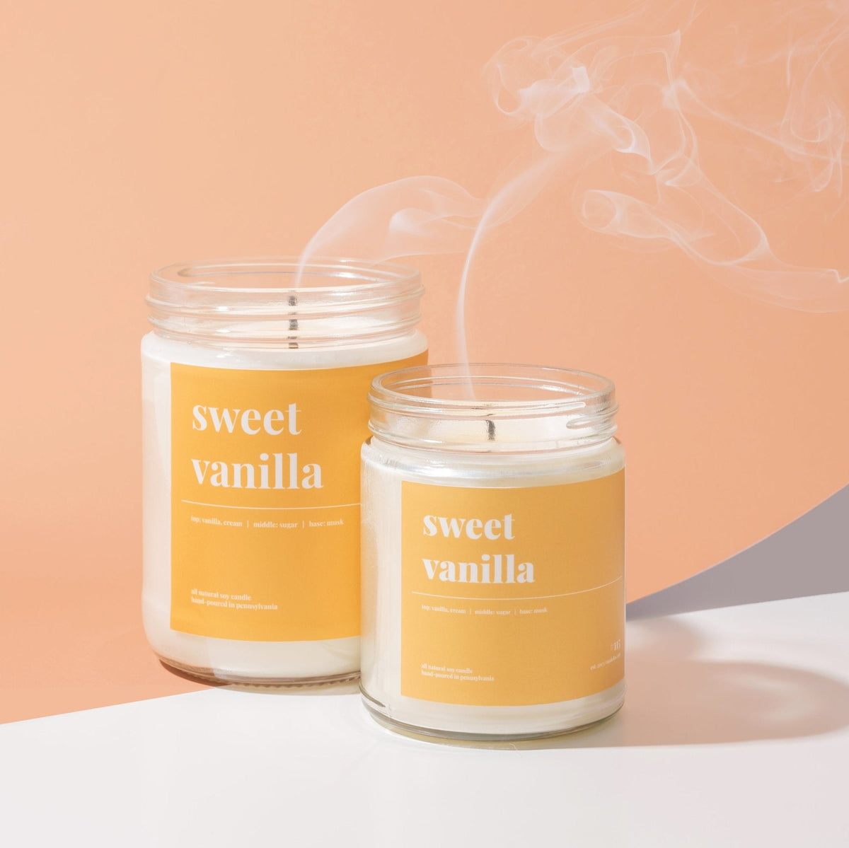 Two sweet vanilla soy candles on a white ledge, one freshly extinguished with a curl of smoke rising, on peach