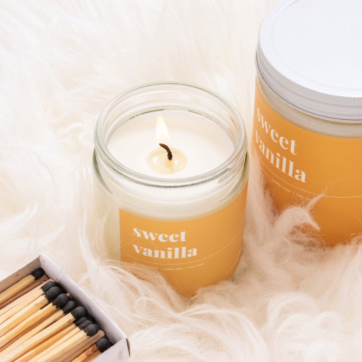 Lit sweet vanilla soy candle with golden label next to a lidded jar and a box of matches on white faux fur