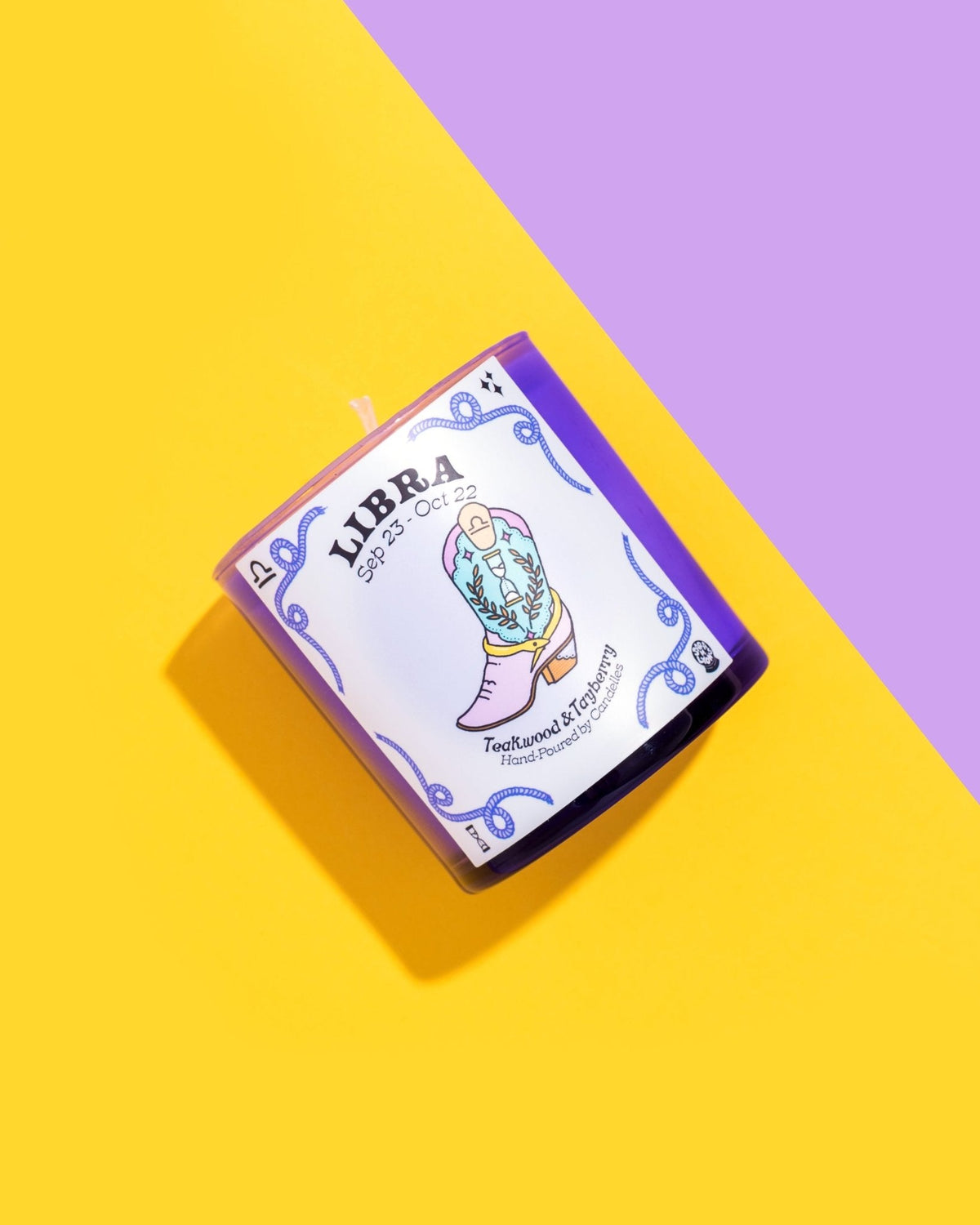Libra teakwood and tayberry zodiac candle tilted across yellow and purple paper with an illustrated boot label