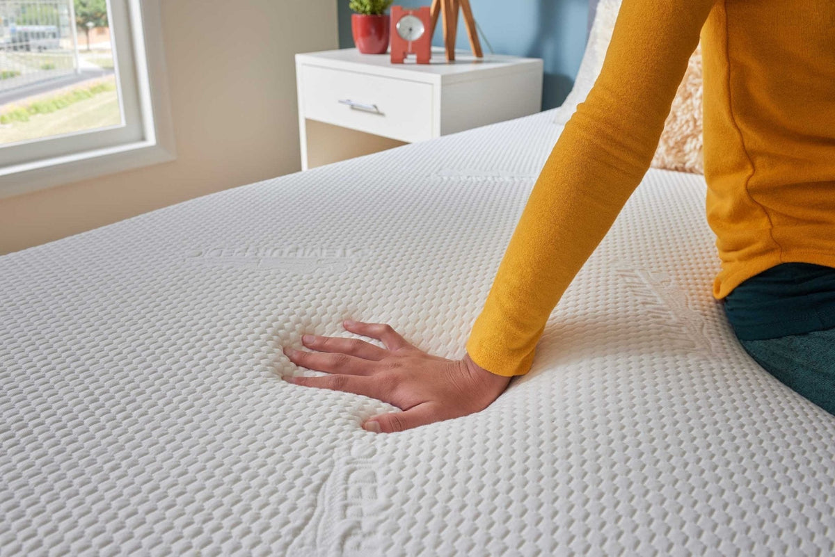 Close-up of a hand sinking into the white topper on a bed in a blue bedroom, showing how the foam conforms