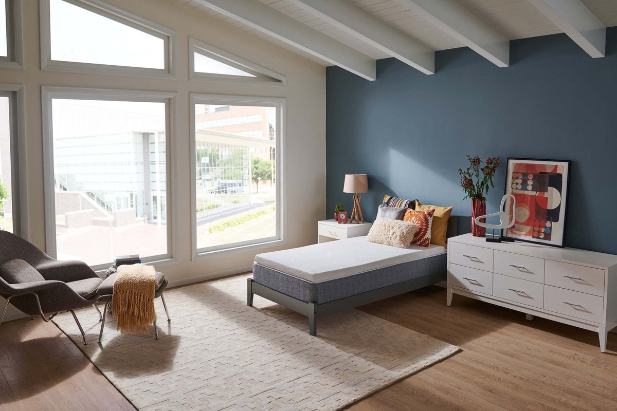 Airy bedroom with a twin bed topped in white foam, corner windows, woven rug and mid-century lounge chair