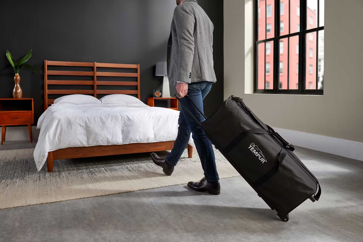 Man wheeling the black travel duffel across a bedroom with a wood platform bed and white bedding behind him