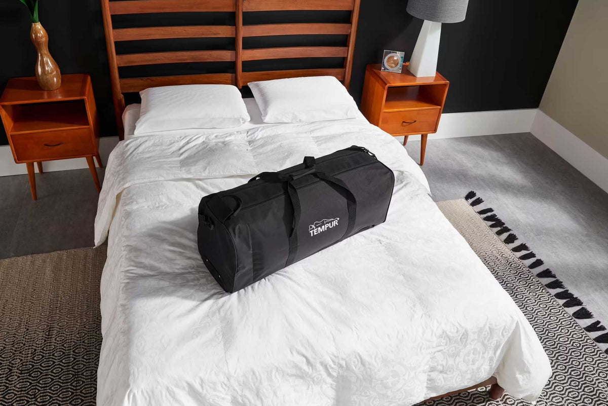 Black wheeled travel duffel bag packed and sitting on a white bed in a bedroom with wood nightstands