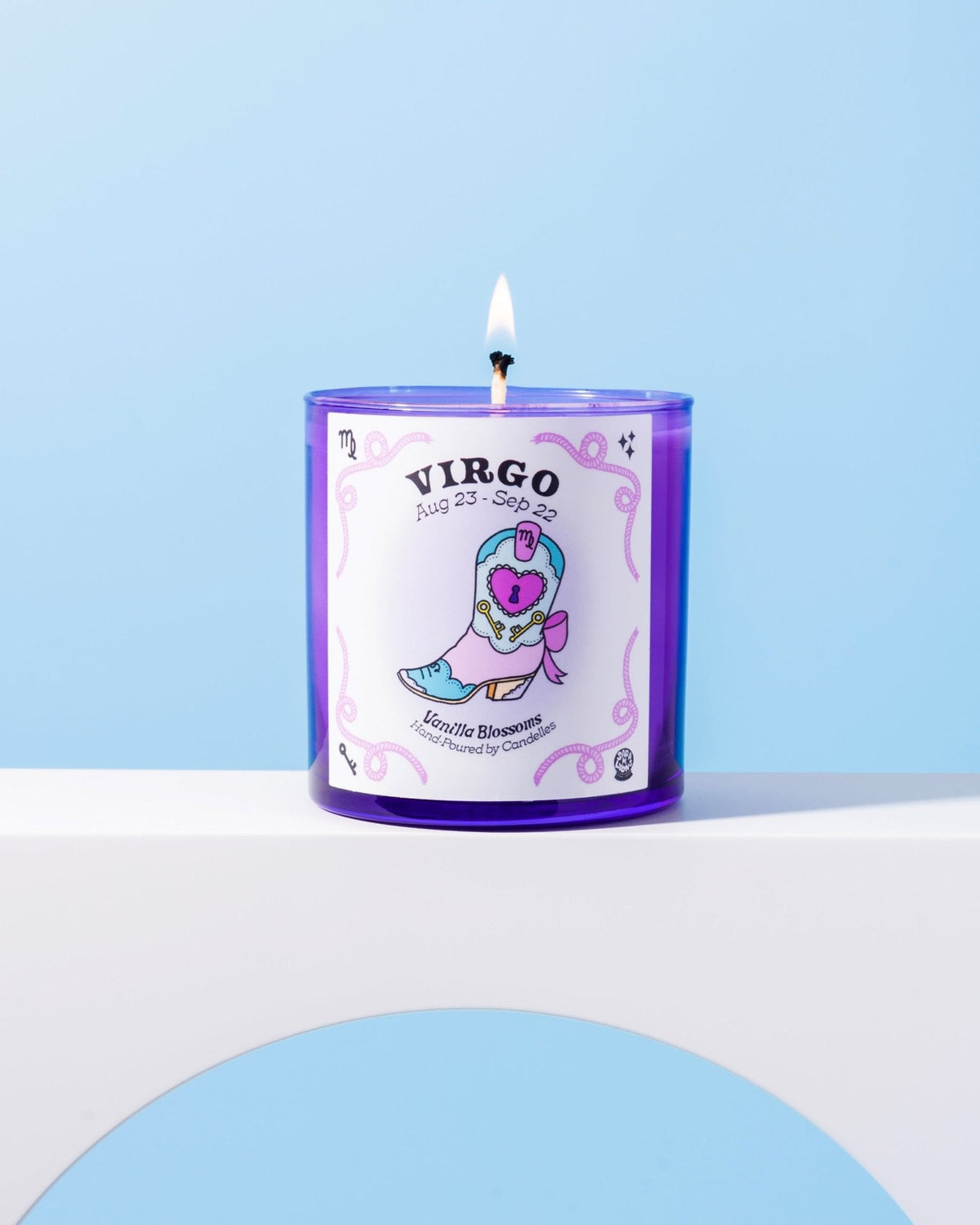 Vanilla blossoms Virgo zodiac candle burning in a purple glass tumbler on a white ledge against pale blue