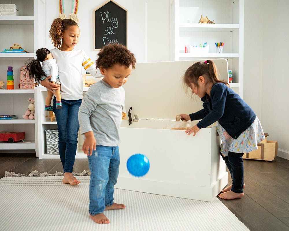 Three children playing beside an open white wooden toy box with a soft-close lid in a bright playroom