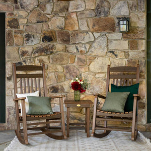 Two brown poly rocking chairs with sage pillows and a small side table with flowers against a stone wall
