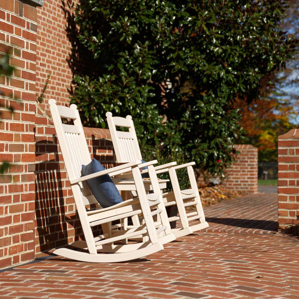Pair of white poly rocking chairs with a blue pillow and side table tucked along a sunny brick walkway