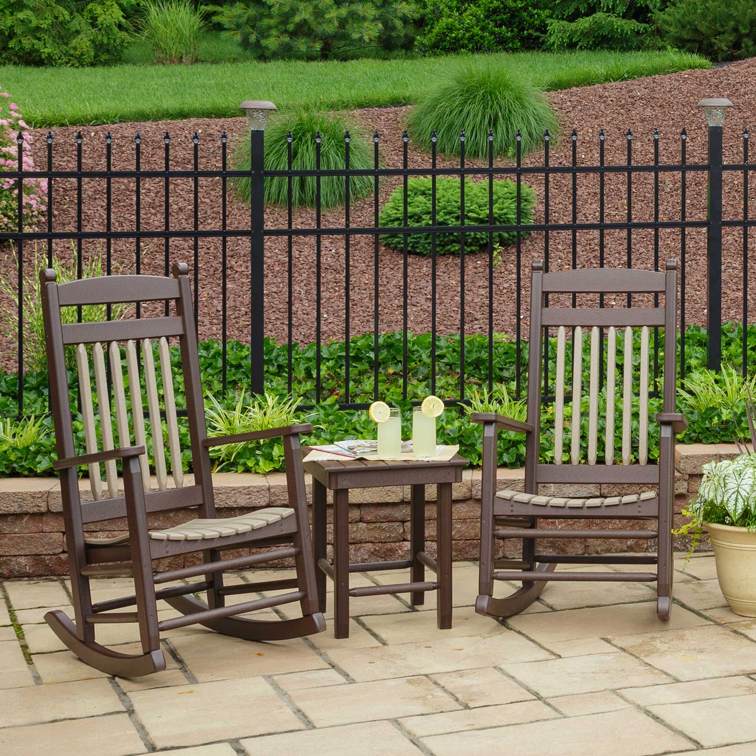 Two brown poly rocking chairs with tan seats flanking a small side table with lemonade on a paver patio