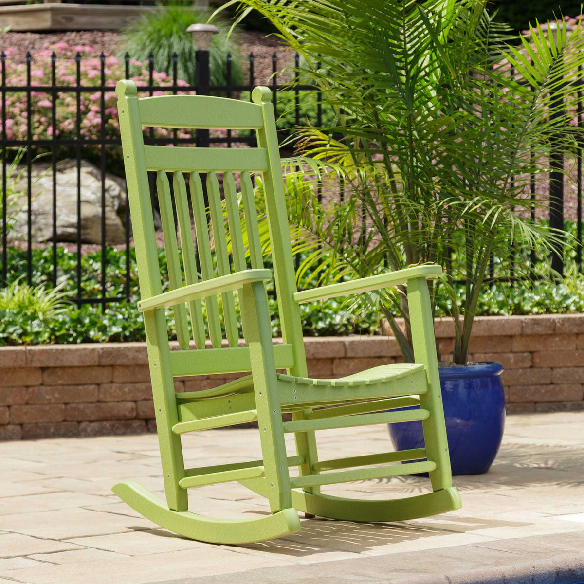 Lime green poly rocking chair with slat back and curved seat beside a blue planter on a paver patio