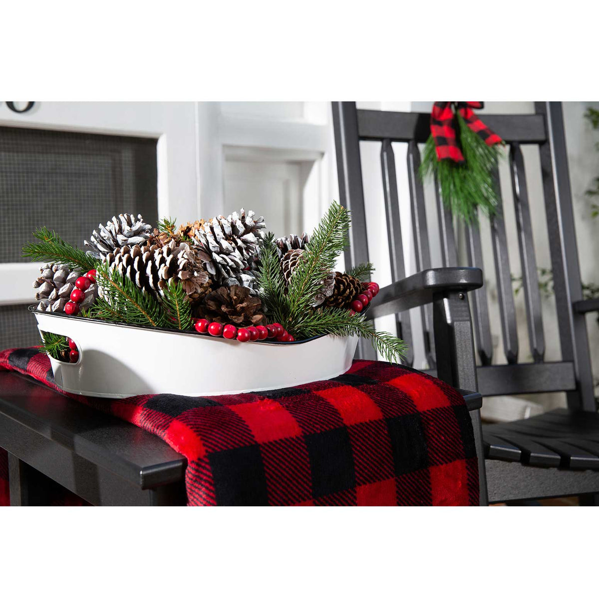 White tray of frosted pinecones and greenery on a black poly side table with a red plaid throw