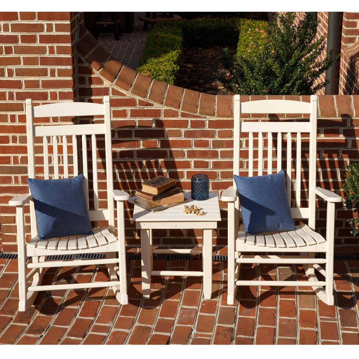 Two white poly rockers with navy pillows beside a small table holding books and a jar on brick pavers