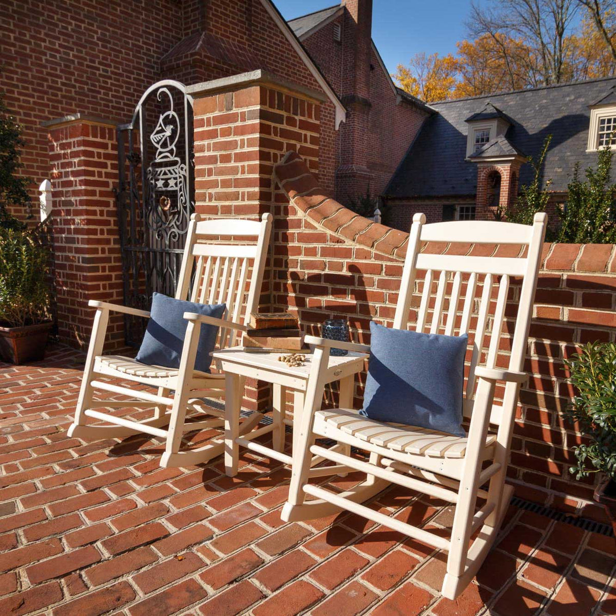 Two cream poly rocking chairs with denim pillows flanking a matching side table on a brick courtyard