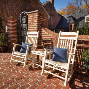 Two cream poly rocking chairs with denim pillows flanking a matching side table on a brick courtyard