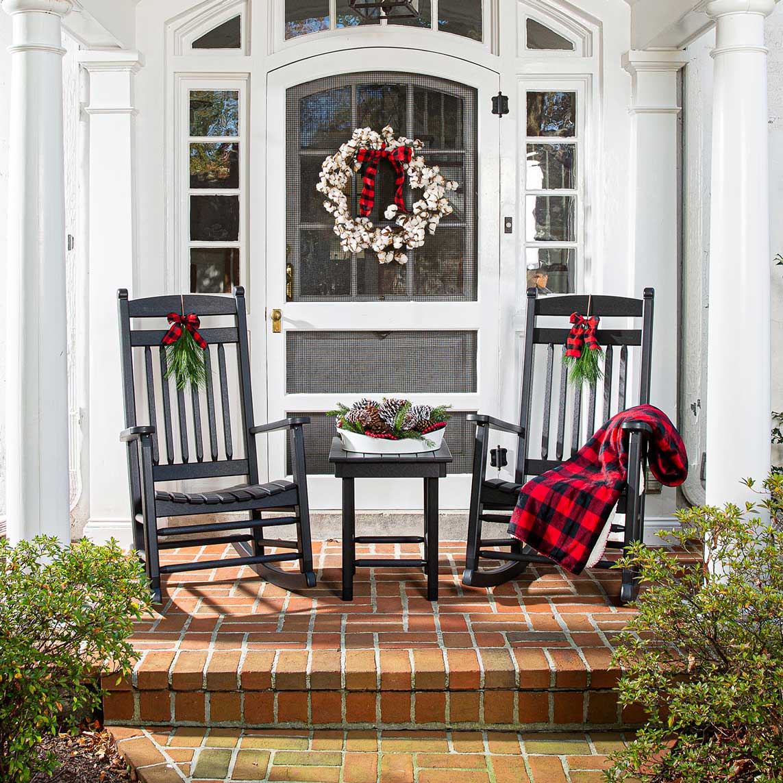 Two black poly rocking chairs with holiday bows, a red plaid throw and a pinecone tray on a white porch