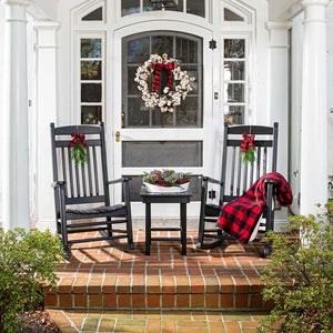 Two black poly rocking chairs with holiday bows, a red plaid throw and a pinecone tray on a white porch